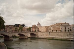Rome, Italy skyline along the Tiber River, with St. Peter's Basilica dome rising above the historic buildings and stone bridge