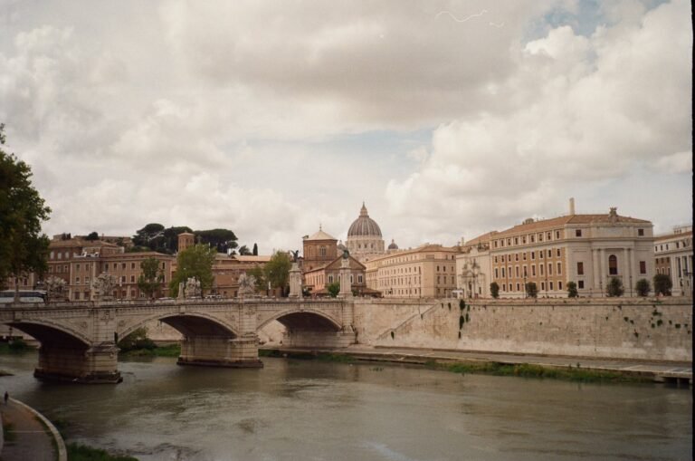 Rome, Italy skyline along the Tiber River, with St. Peter's Basilica dome rising above the historic buildings and stone bridge