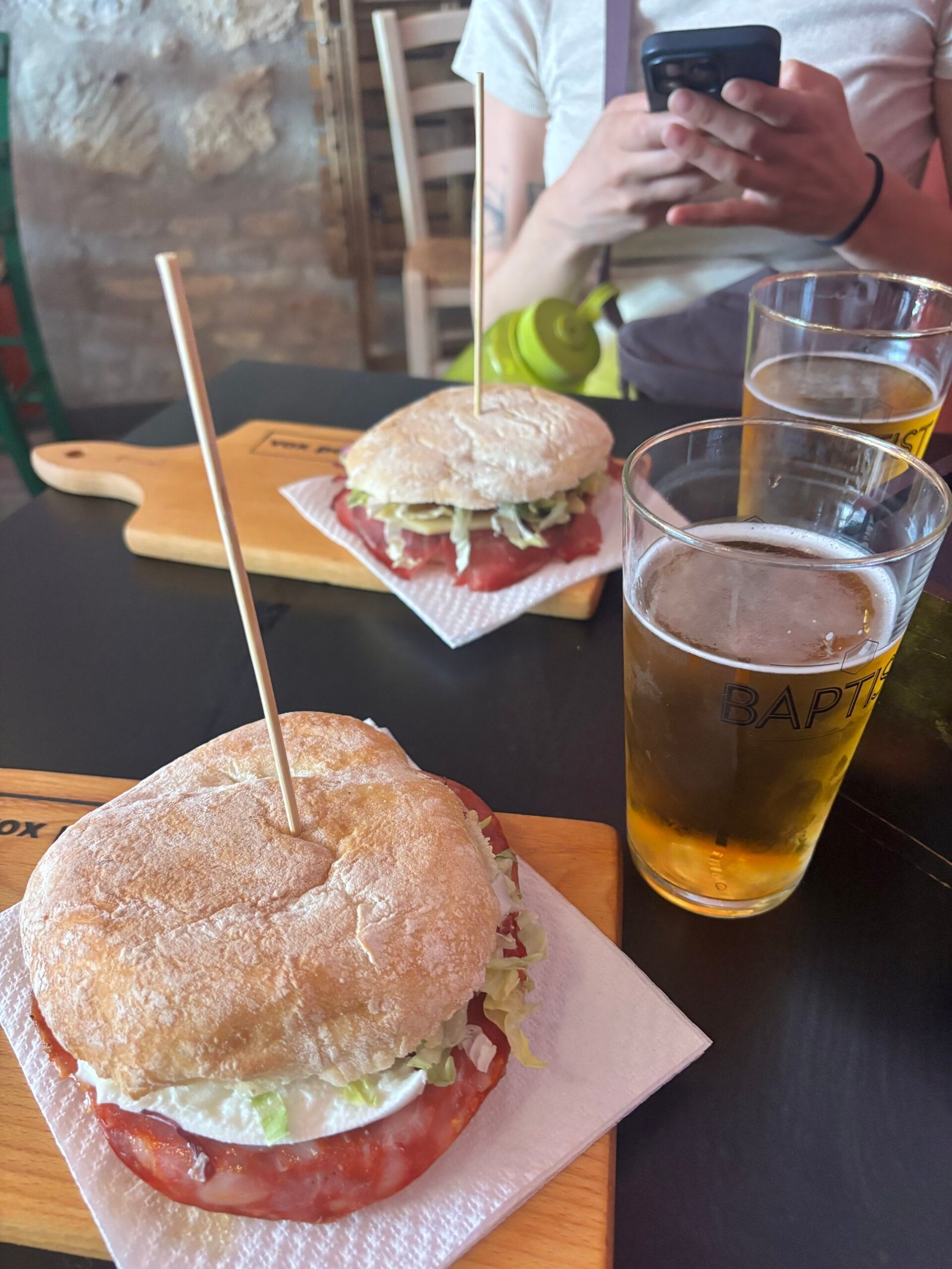 Fresh sandwiches loaded with prosciutto, cheese, and lettuce on crusty rolls, served on wooden boards with two glasses of cold beer, a casual outdoor lunch in Rome